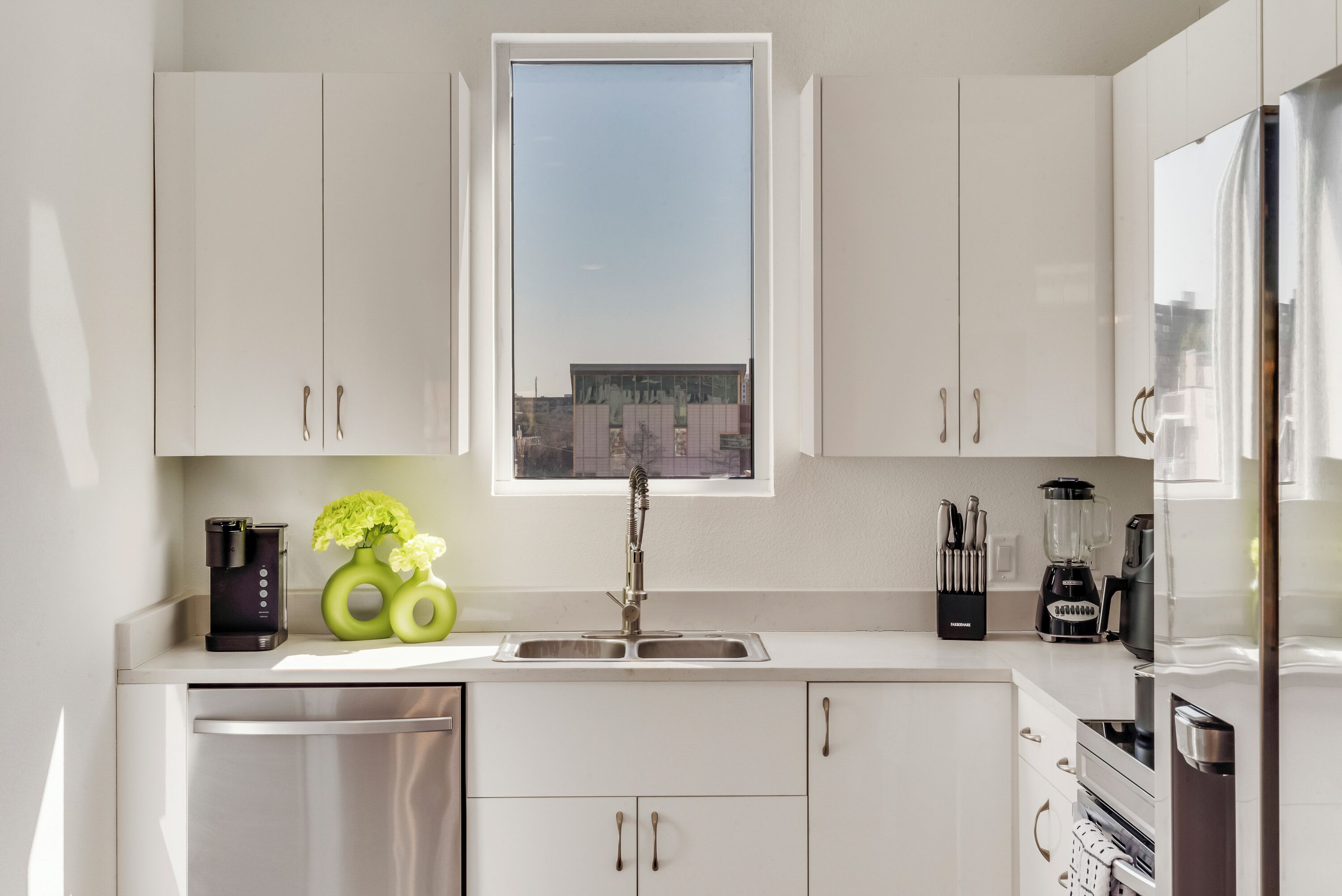 A modern kitchen with white cabinets, a stainless steel dishwasher, a window with a city view, and various kitchen appliances.