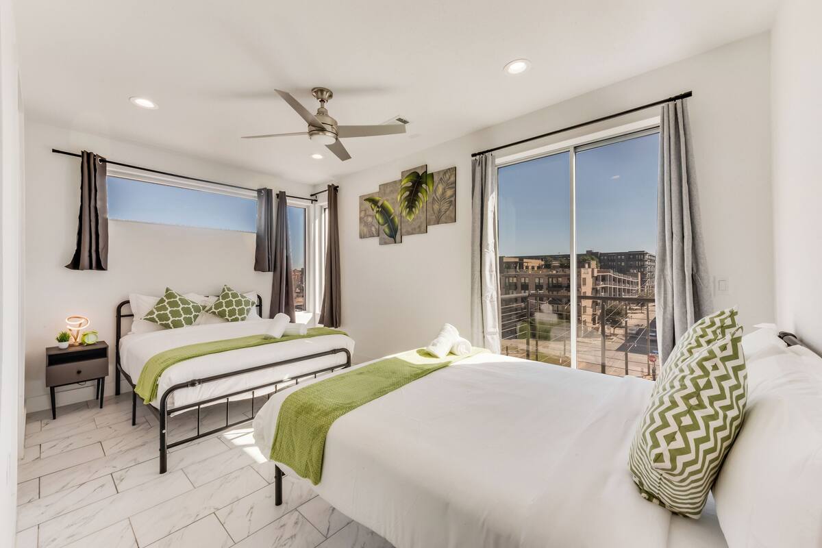 Quarto de hotel moderno com duas camas, ventilador de teto, janelas amplas com cortinas e vista para a cidade.