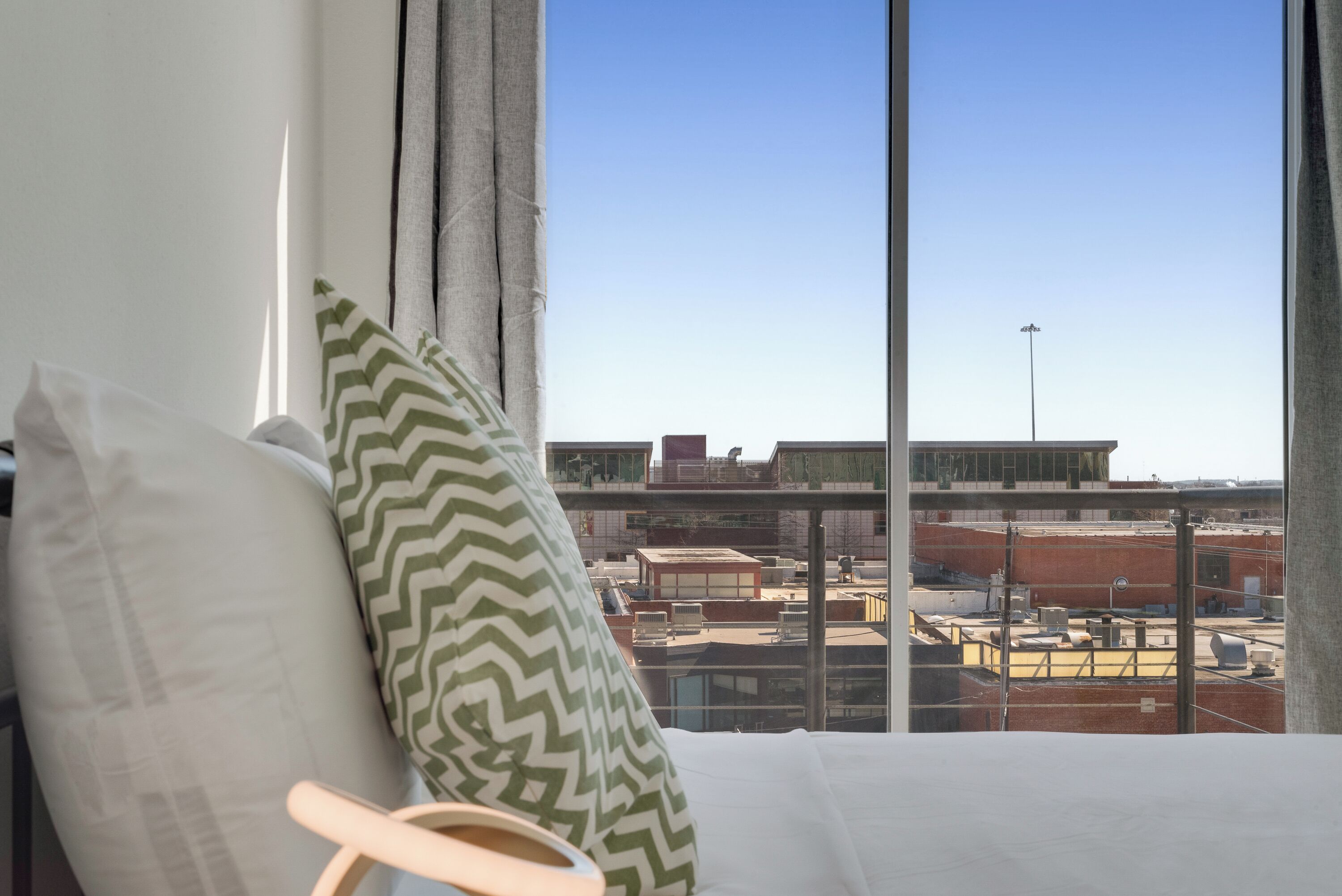 A hotel room with a large window offering a view of the cityscape, a bed with white bedding and a green and white patterned pillow, and a wooden chair.