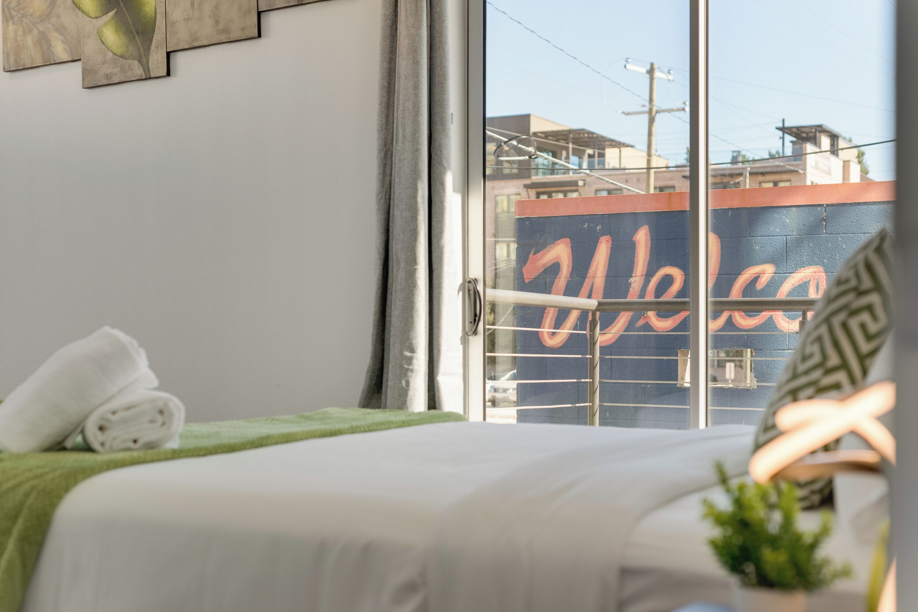 A hotel room with a bed, a green towel, a plant, and a view of the outside through a window with a 'Welcome' sign.