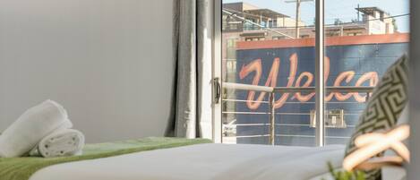 A hotel room with a bed, a green towel, a plant, and a view of the outside through a window with a 'Welcome' sign.