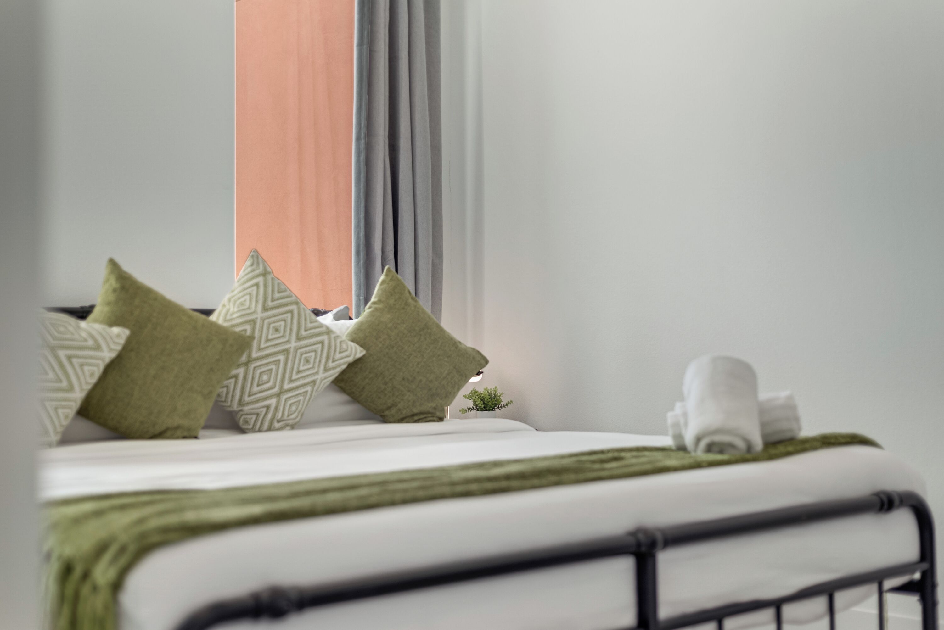 A neatly made bed with green and patterned pillows, a rolled white towel, and a small plant on the nightstand.
