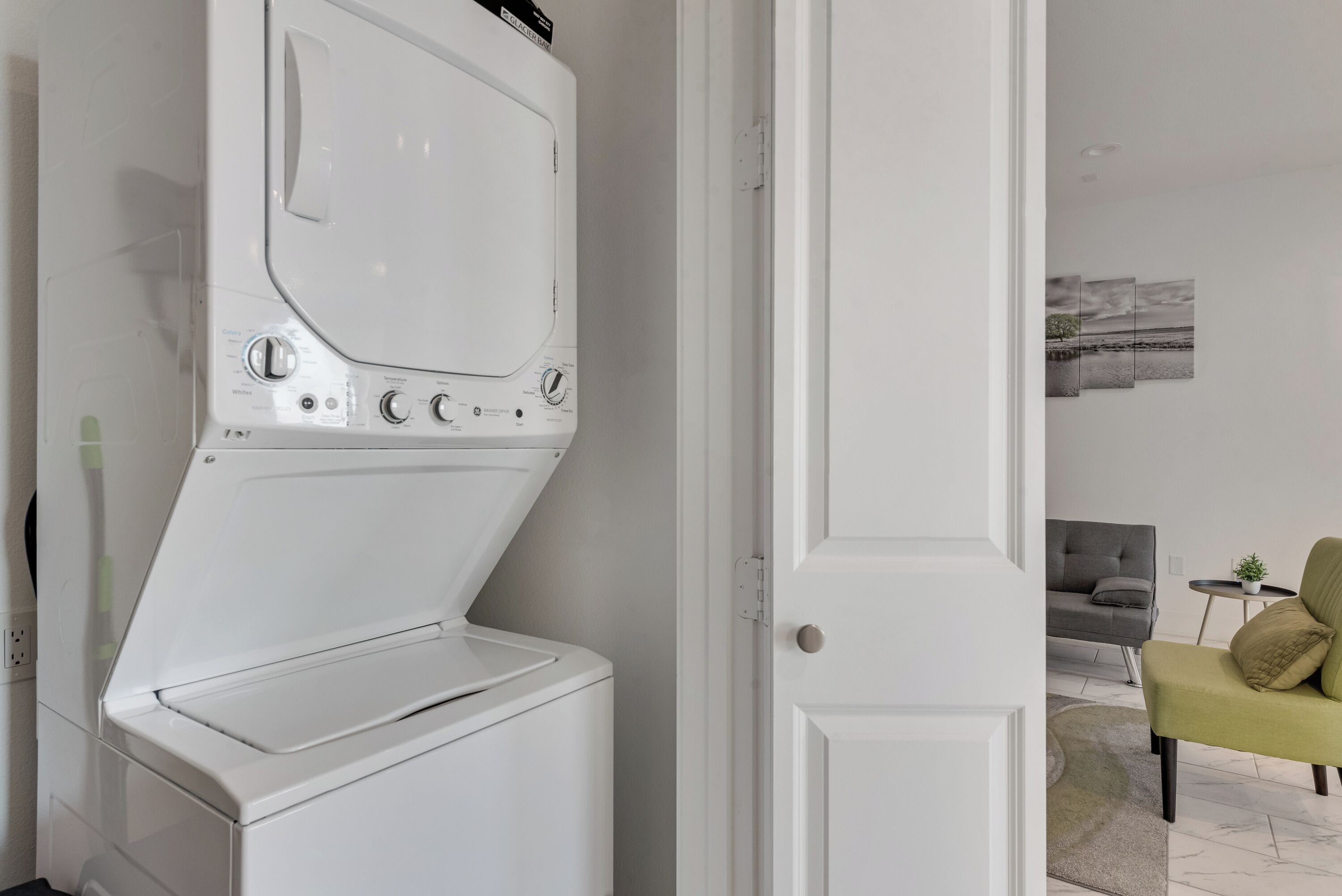 A modern laundry room with a front-loading washer and dryer, a white door, and a living area with a gray sofa and a green armchair.