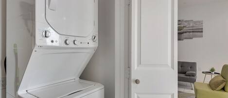 A modern laundry room with a front-loading washer and dryer, a white door, and a living area with a gray sofa and a green armchair.