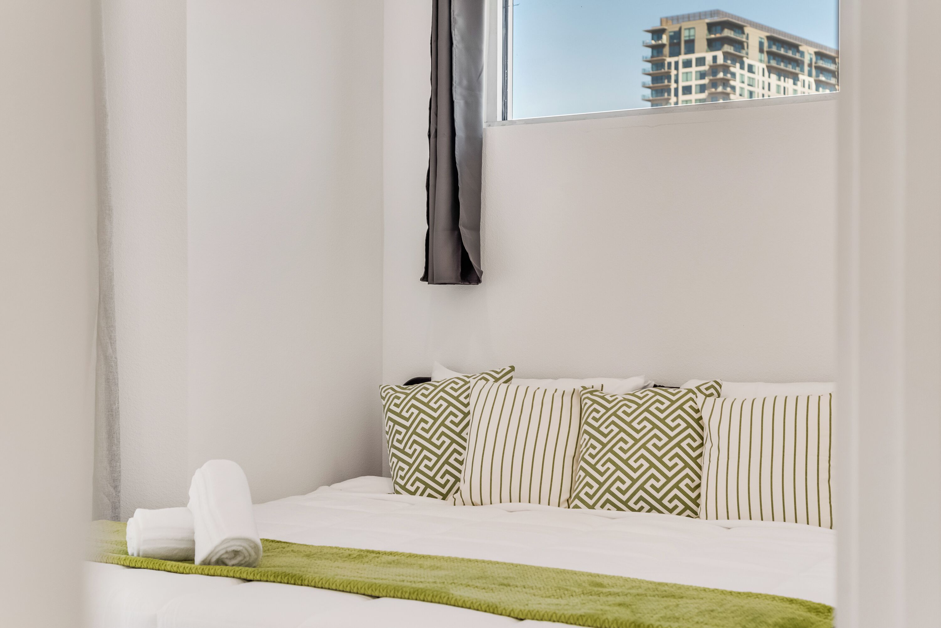 Une chambre d’hôtel avec un lit, des oreillers, un plaid vert et une fenêtre donnant sur un immeuble.