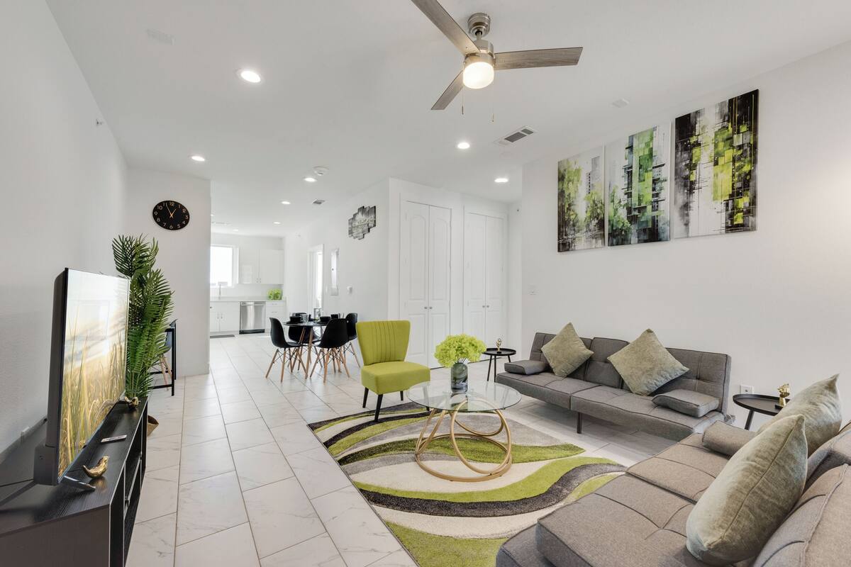 Uma sala de estar moderna com um sofá cinzento, uma poltrona verde, uma mesa de centro de vidro e um tapete decorativo. Há um ventilador de teto, um quadro na parede e uma área de jantar ao fundo.