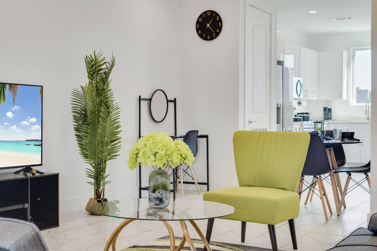 Uma sala de estar moderna com uma mesa de centro de vidro, uma poltrona verde, uma grande planta em vaso, uma televisão de tela plana e uma cozinha ao fundo.