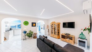 Un salon moderne avec un canapé, un téléviseur et un meuble en bois. On peut apercevoir une cuisine à travers une arche.
