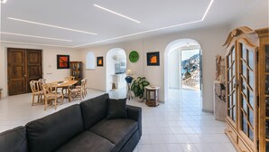 Un vaste espace de vie avec un canapé foncé, une table à manger en bois et des chaises. Les murs sont ornés d’œuvres d’art encadrées, et on y trouve également une plante en pot. Une arche ouverte offre une vue sur une ville côtière.