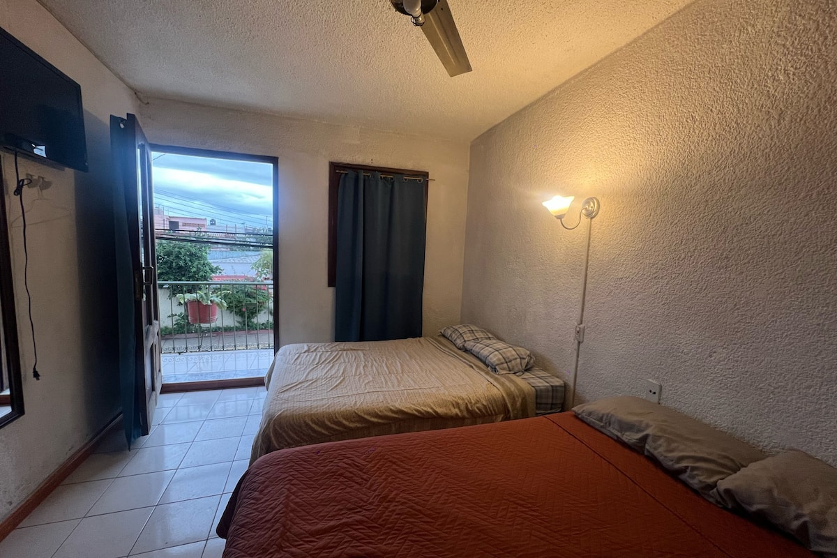 Une chambre avec deux lits, un ventilateur de plafond, une fenêtre avec des rideaux et un luminaire fixé au mur.