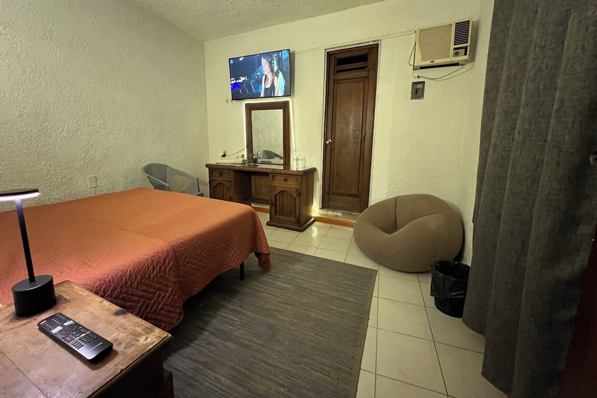 A bedroom with a bed, a desk, a chair, a television, a wardrobe, and an air conditioning unit.