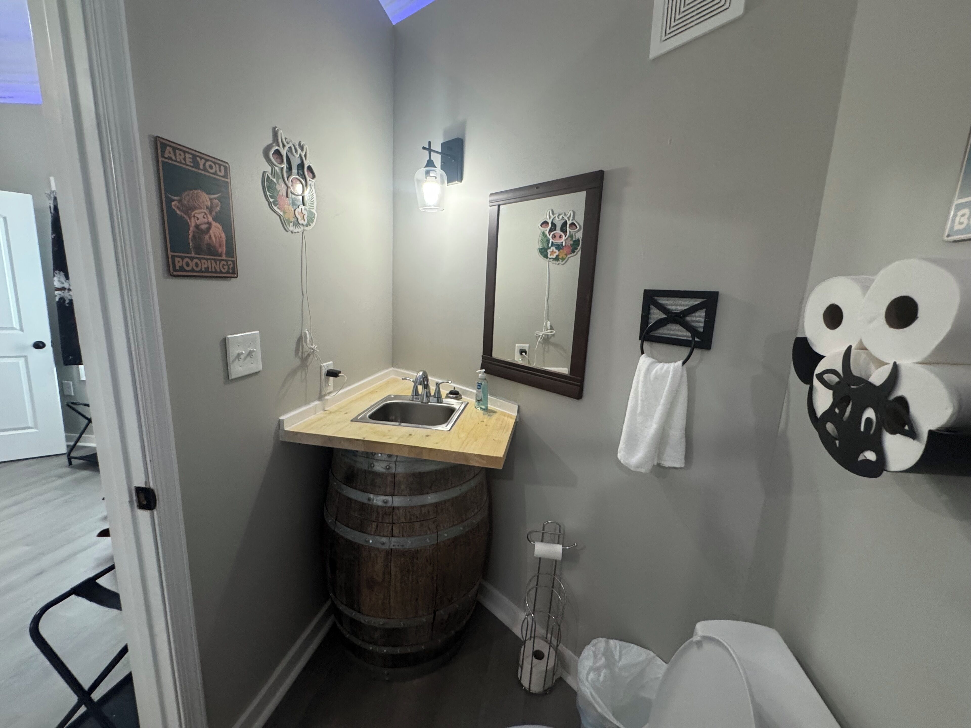 A bathroom with a wooden vanity, a sink, a mirror, and a toilet. There are decorative wall hangings and a towel rack.