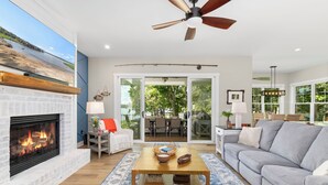 A living room with a fireplace, a ceiling fan, and a large window overlooking a deck. - New! Lakefront 4BR, Kayaks, Paddleboards, Games (Littleton)