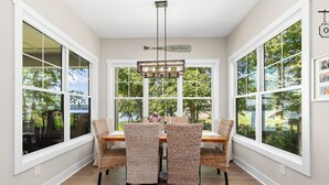 A dining area with a table and four wicker chairs, large windows with white frames, and a hanging light fixture. - New! Lakefront 4BR, Kayaks, Paddleboards, Games (Littleton)