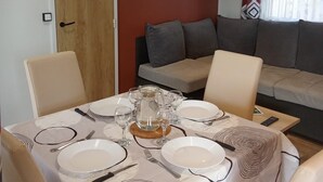 Uma sala de jantar com mesa posta para quatro pessoas, um sofá e uma pequena estante com livros e um laptop.