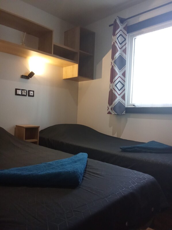 A room with two beds, a small wooden shelf, a wall-mounted light, and a window with patterned curtains.