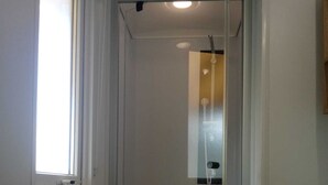 Um espaço de chuveiro pequeno e fechado, com porta de vidro, janela lateral e radiador branco na parte inferior.