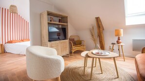 A modern living room with a television on a wooden cabinet, a wicker chair, a round table with a lamp, and a bed with striped curtains. - Cosy apartment in the heart of Angers (Angers)