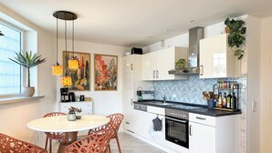 A modern kitchen with white cabinets, a black countertop, and a built-in oven. There is a dining area with a round table and wicker chairs. - Hejm Mountain Vibe & Design vacation apartment - 2 min to the bike park and ski slope (Winterberg)