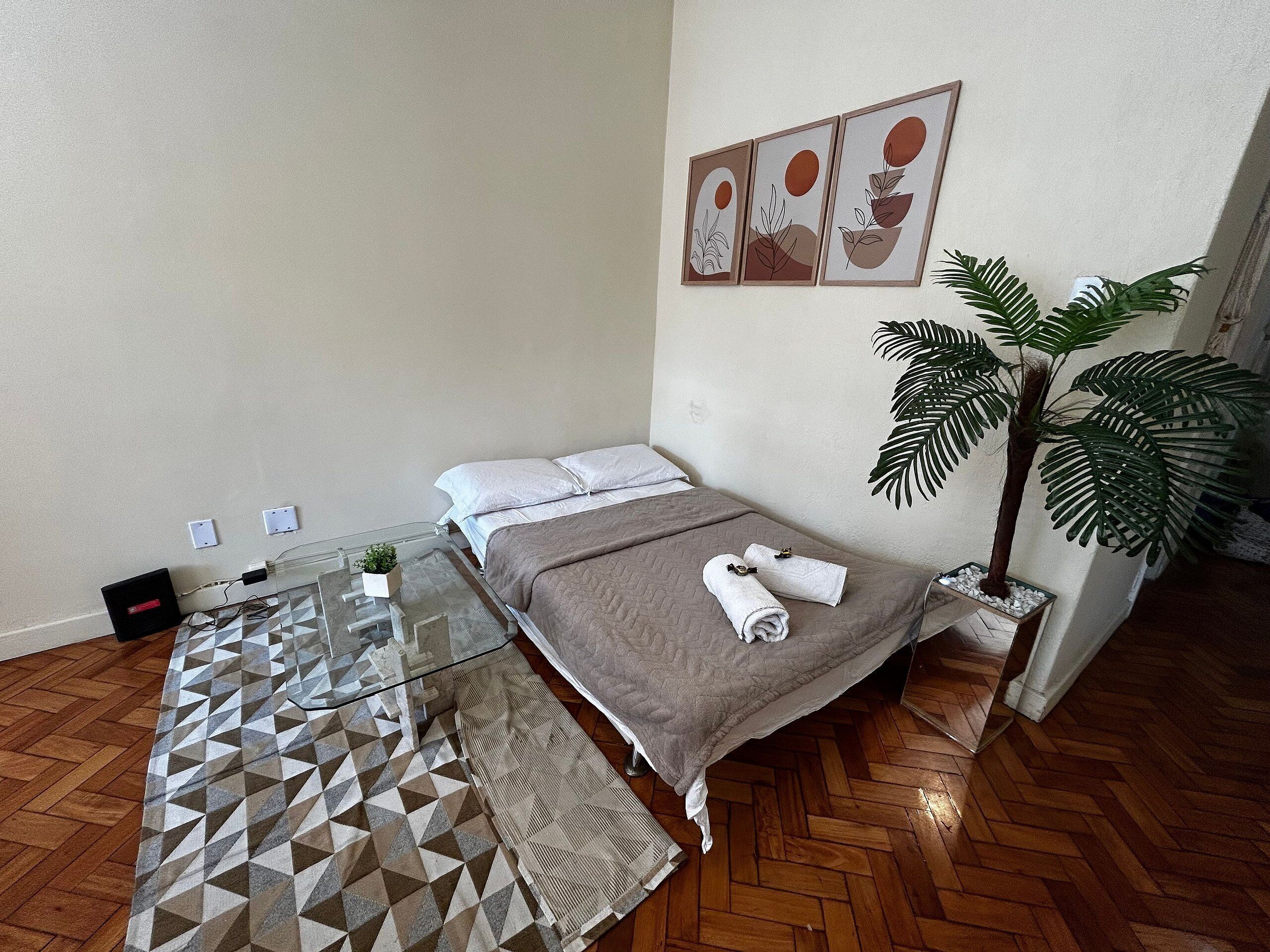 Een moderne slaapkamer met een bed, een glazen tafel met een plant, een grote kamerplant en een geometrisch vloerkleed.
