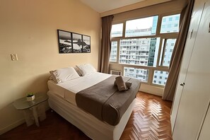 Ein Schlafzimmer mit einem Bett, einem Nachttisch, einer Topfpflanze, einem Fenster mit Blick auf die Stadt und einer Tür.