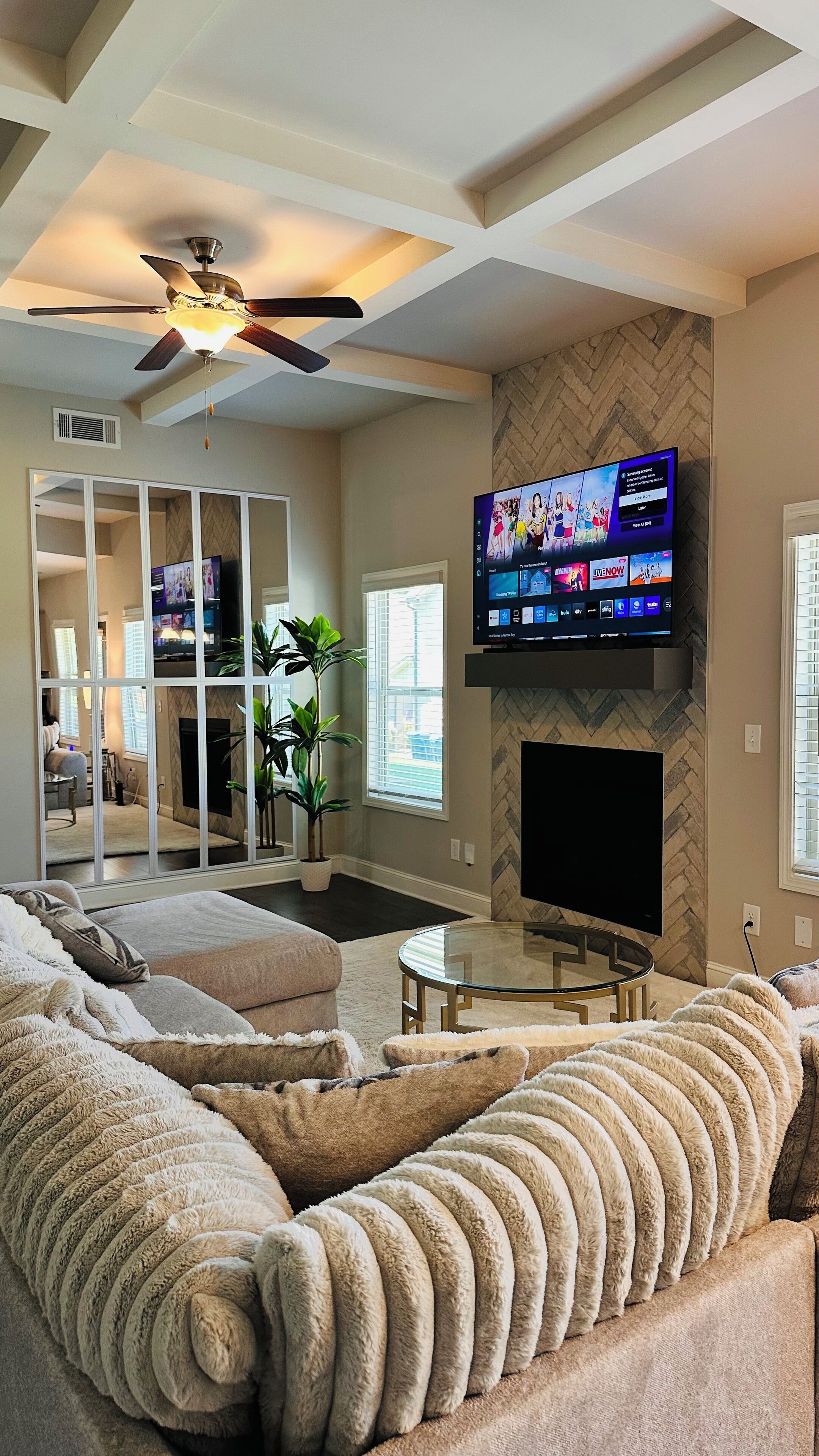 A living room with a large flat-screen TV mounted on the wall, a ceiling fan, a fireplace, and a cozy sectional sofa.
