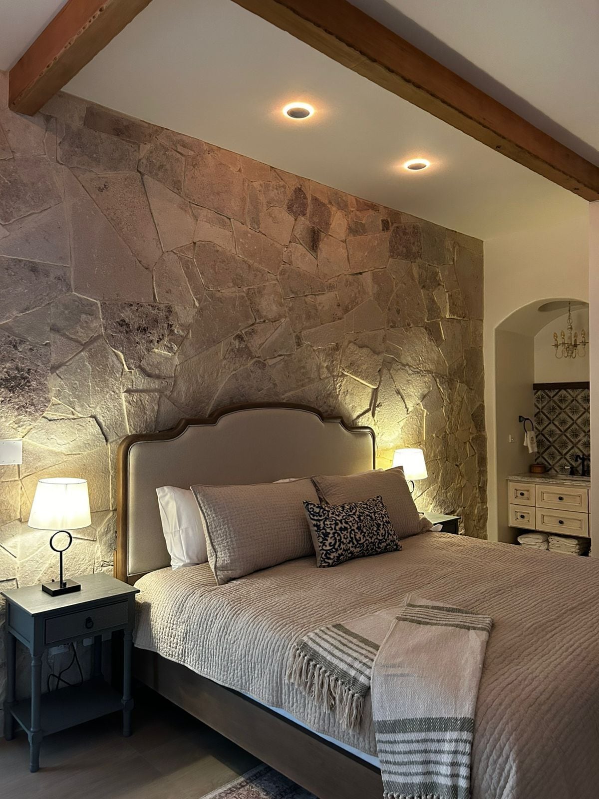A bedroom with a stone wall, a bed with a headboard, two bedside tables with lamps, and a small bathroom with a mirror and towel rack.