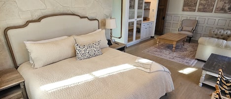 Une chambre dotée d’un grand lit, d’une tête de lit en bois, d’un mur en pierre et offrant une vue sur un espace de vie comprenant un canapé, un fauteuil et des œuvres d’art.