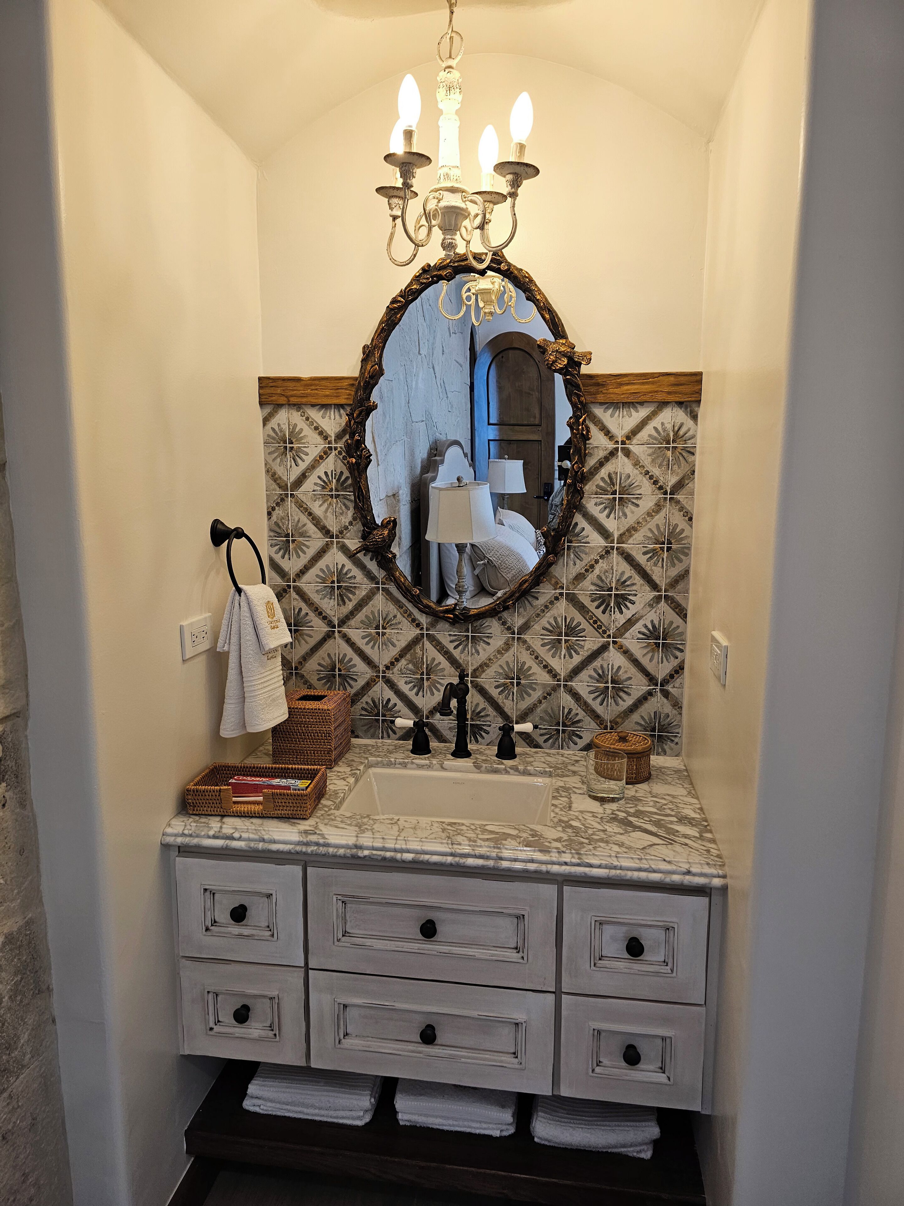 A bathroom with a marble sink, a mirror with an ornate frame, a chandelier, and patterned tiles.