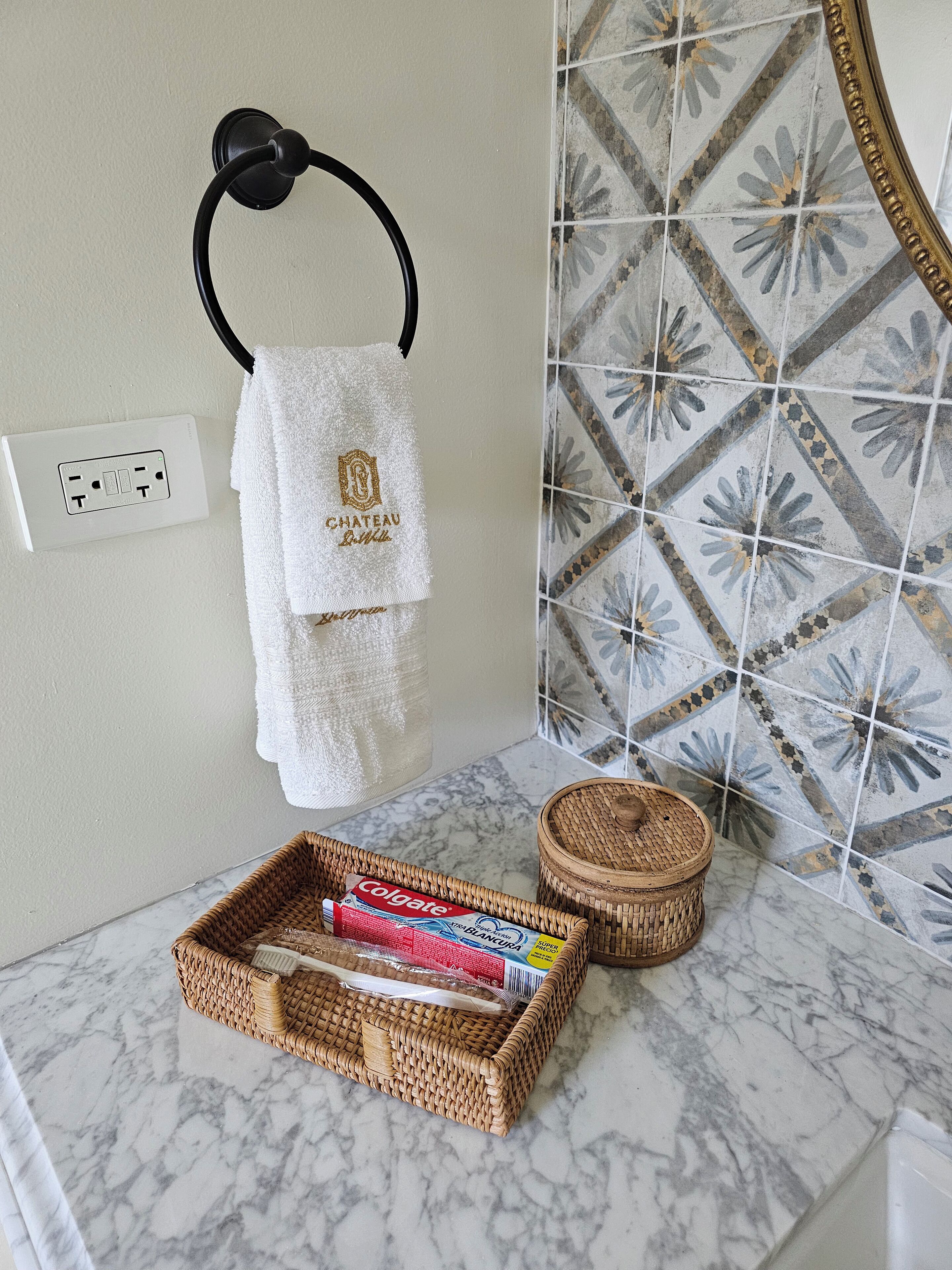 Een badkamer met een witte handdoek aan een zwarte handdoekhouder, een doos Colgate tandpasta en een rieten mandje op een marmeren aanrecht.