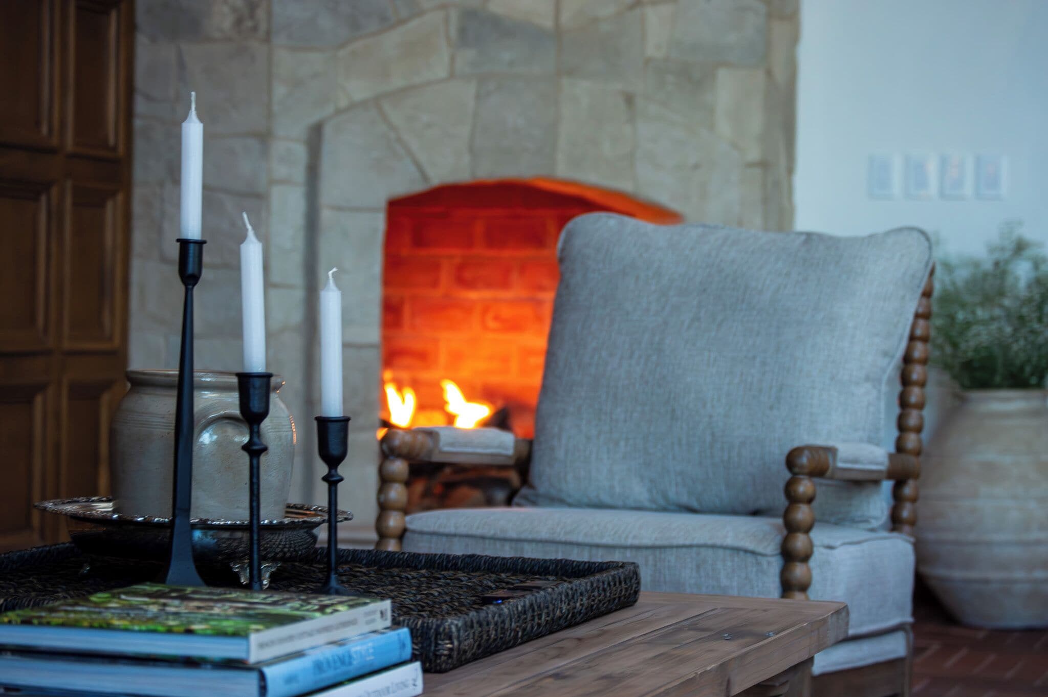 A cozy room with a stone fireplace, a comfortable armchair, and a wooden table with books and candles.