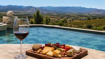 A poolside setting with a glass of red wine, a cheese board with various cheeses, fruits, and nuts, and a view of mountains in the background.