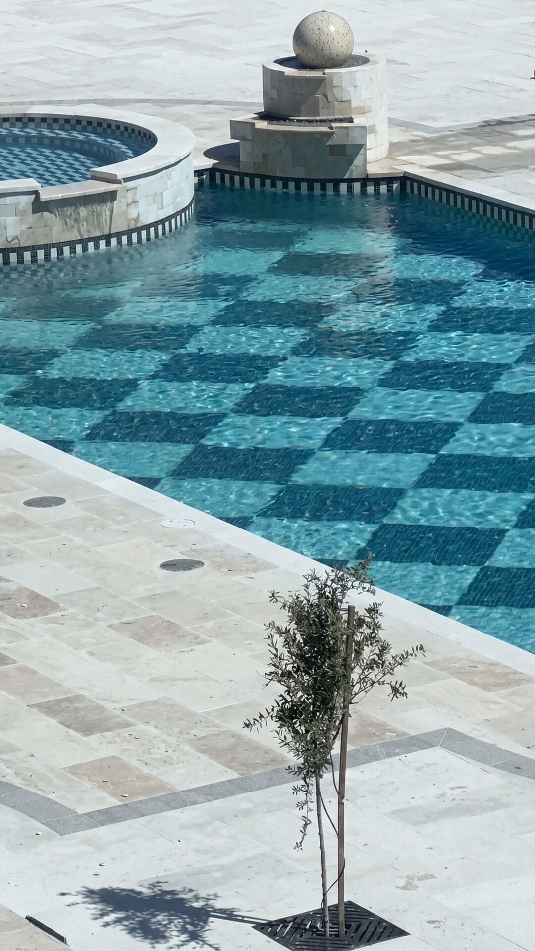 Una piscina con diseño de cuadros, una fuente central y un pequeño árbol en primer plano.