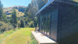A wooden cabin with a deck, surrounded by a grassy area and trees, with a hill in the background.