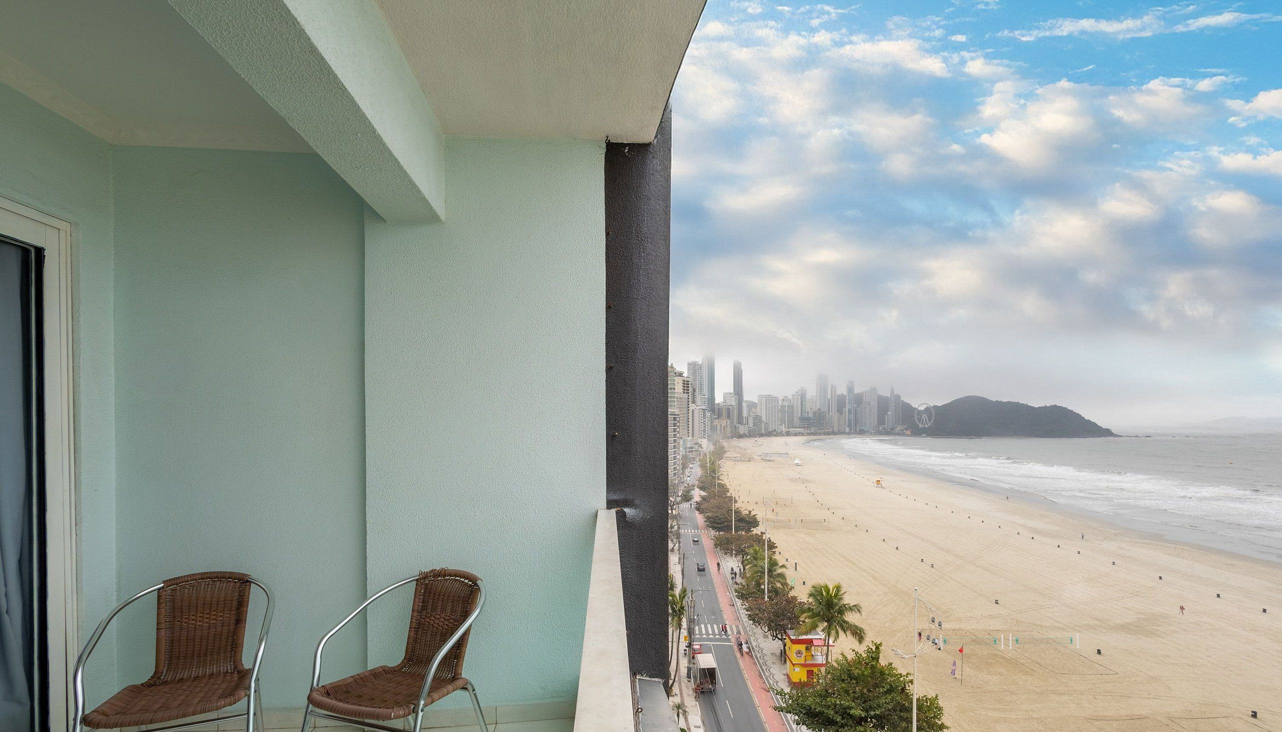A balcony with two wicker chairs, overlooking a beach with a city skyline and a Ferris wheel in the distance.