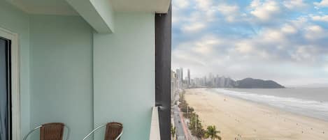 A balcony with two wicker chairs, overlooking a beach with a city skyline and a Ferris wheel in the distance.
