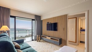A modern hotel room with a large TV mounted on a wooden panel wall, a sofa, a dining table, and a balcony with a view of the sea. - Apt w/ Balcony and Oceanfront View in BC - Ipc0901 (Balneário Camboriú)