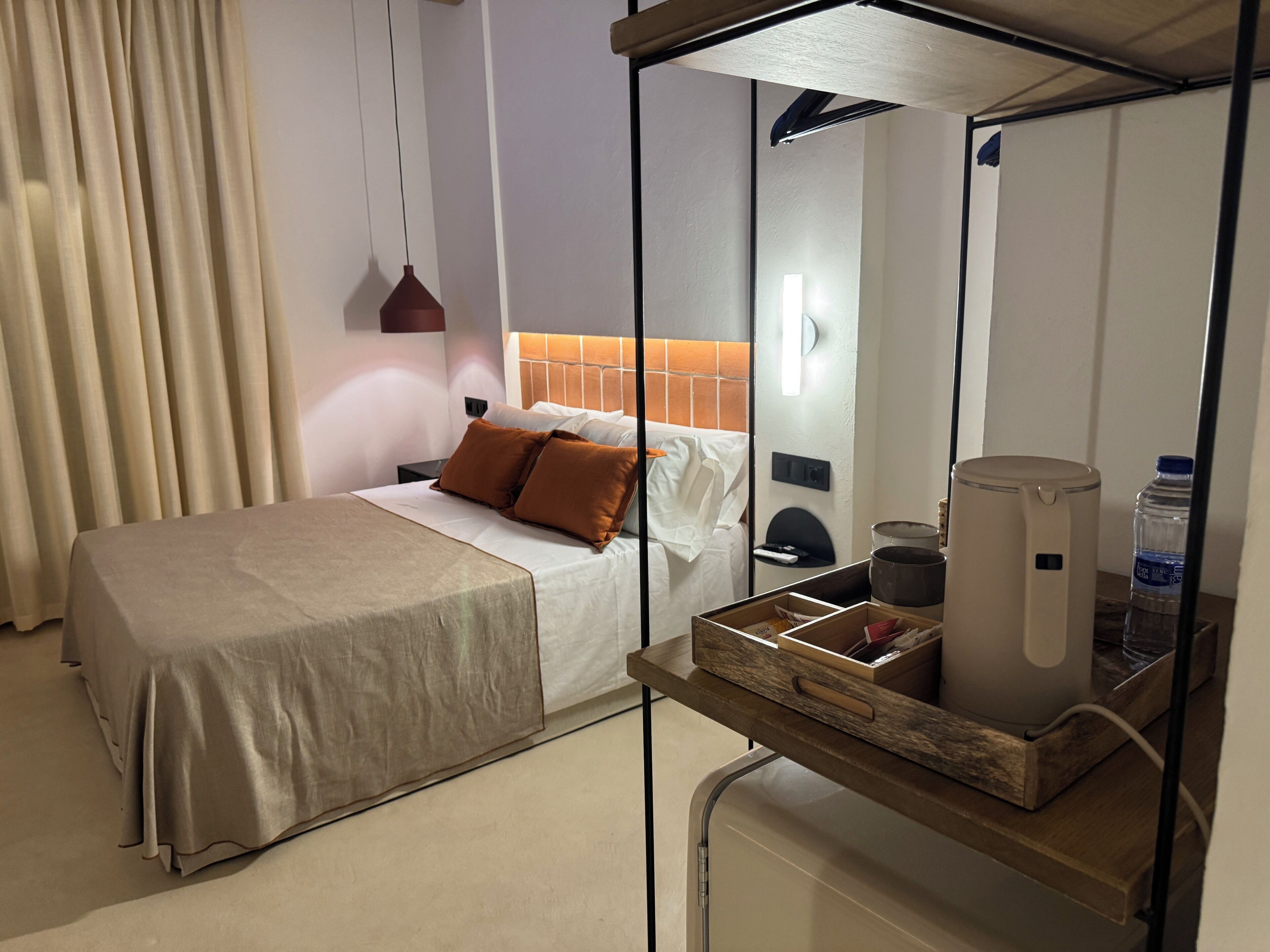 Quarto de hotel bem arrumado com uma cama, uma mesinha de cabeceira com uma chaleira, uma garrafa de água e uma pequena caixa de madeira.