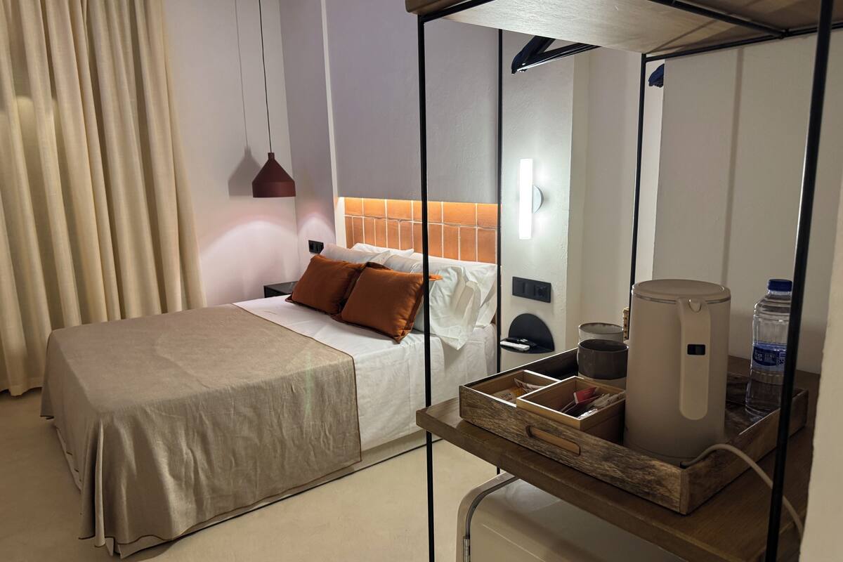 Une chambre d’hôtel bien rangée, avec un lit, une table de chevet sur laquelle se trouvent une bouilloire et une bouteille d’eau, ainsi qu’une petite boîte en bois.