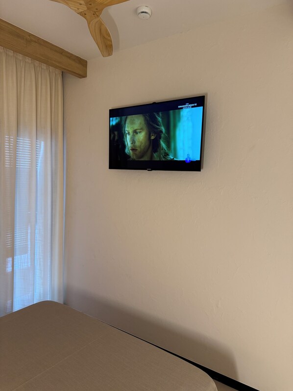 Quarto com televisão na parede, uma cama e janela com cortinas.