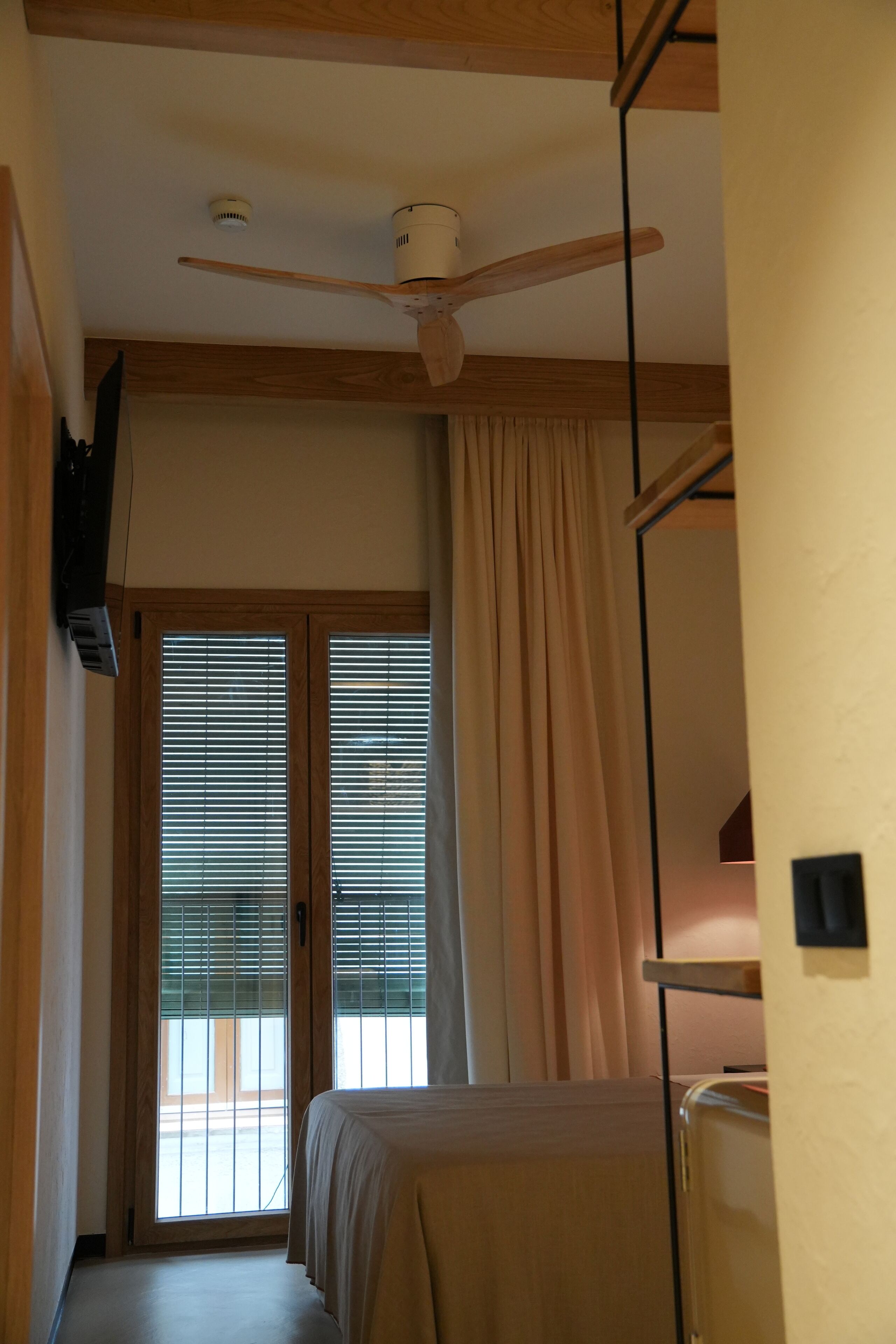 Una habitación con ventilador de techo, una cama con edredón color beige, una ventana con persianas y una puerta con cortina.