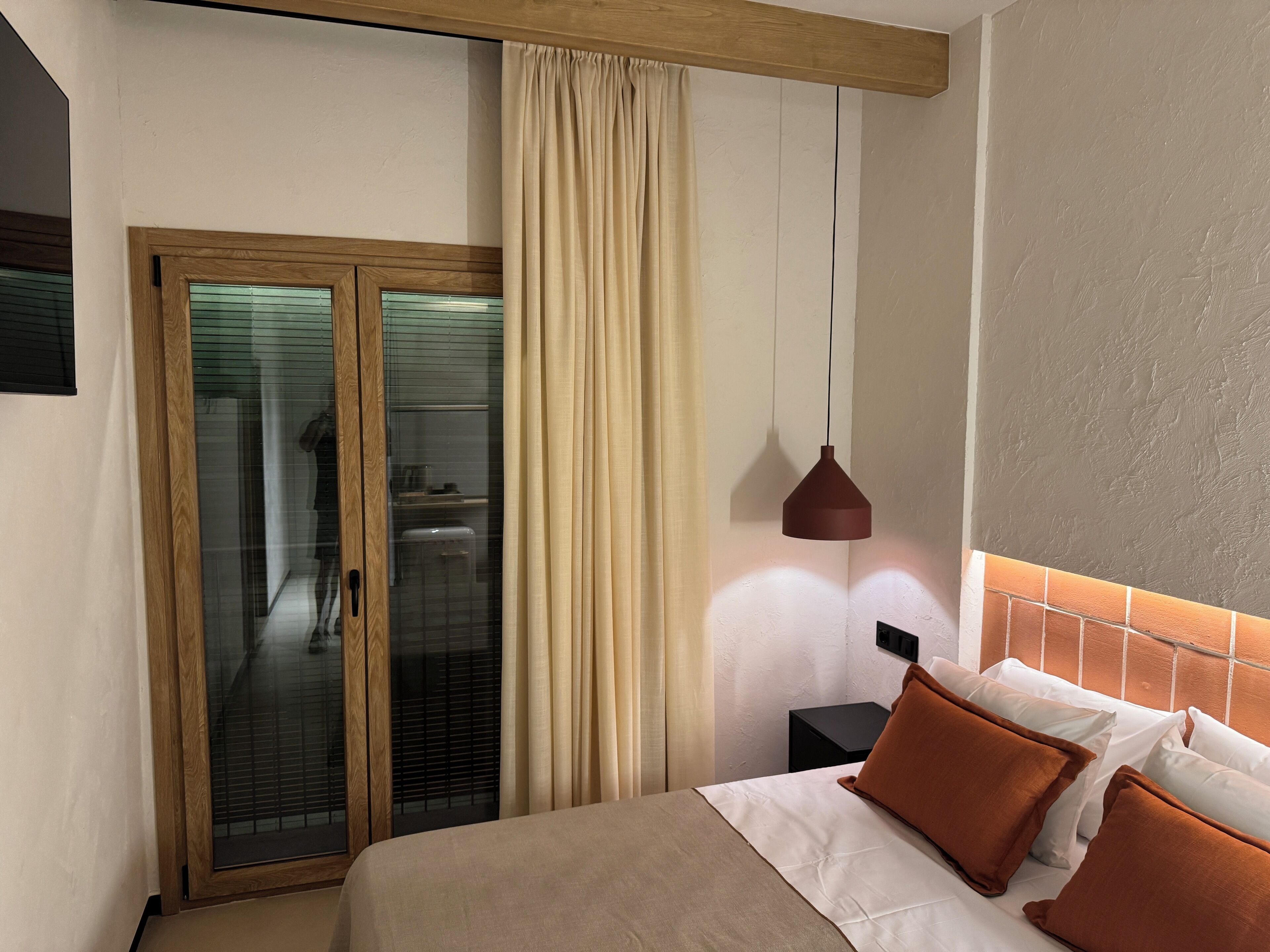 Quarto de hotel com cama, mesa de cabeceira, luminária pendente e porta de vidro deslizante que dá para um banheiro.