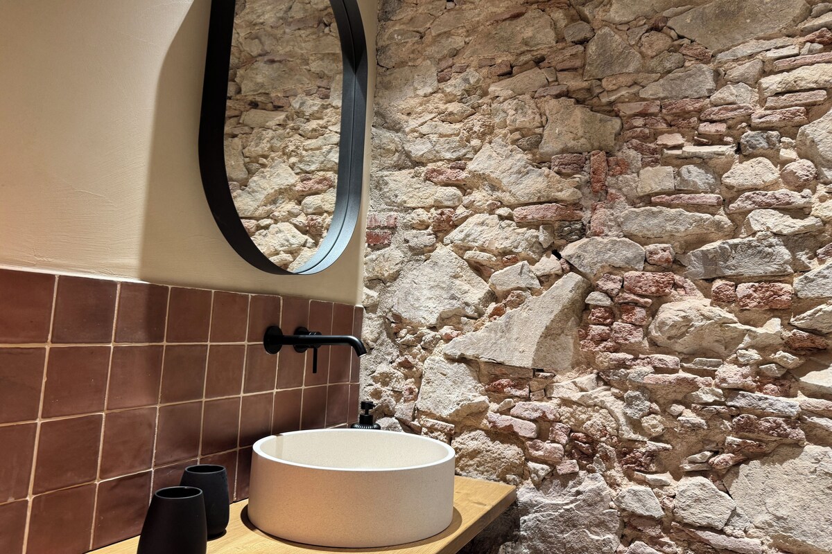 Een badkamer met een stenen muur, een rond wastafel, een spiegel en bruine tegels.