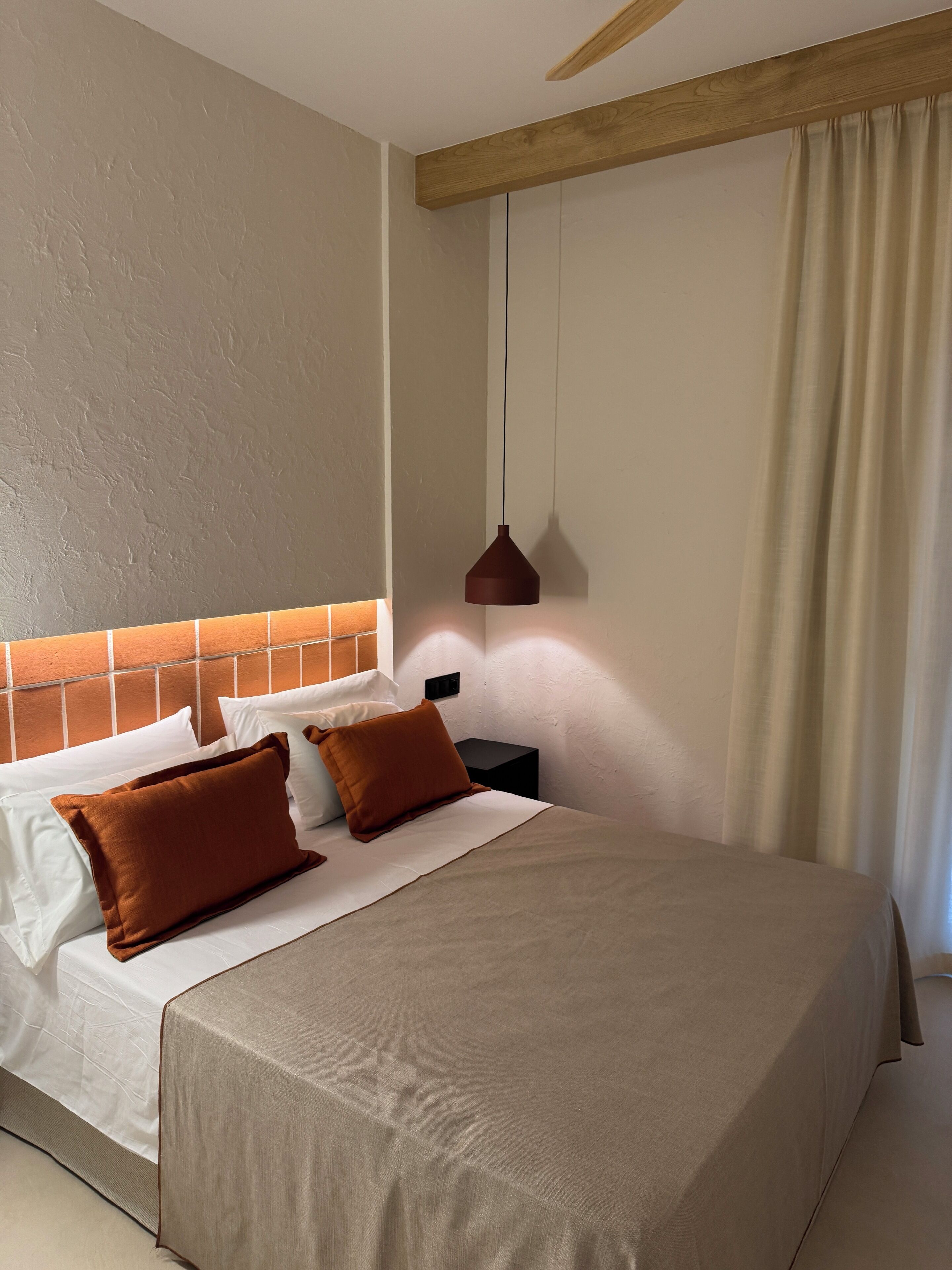 Un letto ben rifatto con copriletto color beige e due cuscini marroni, una testiera in legno, un comodino con una lampada e una finestra con tende.