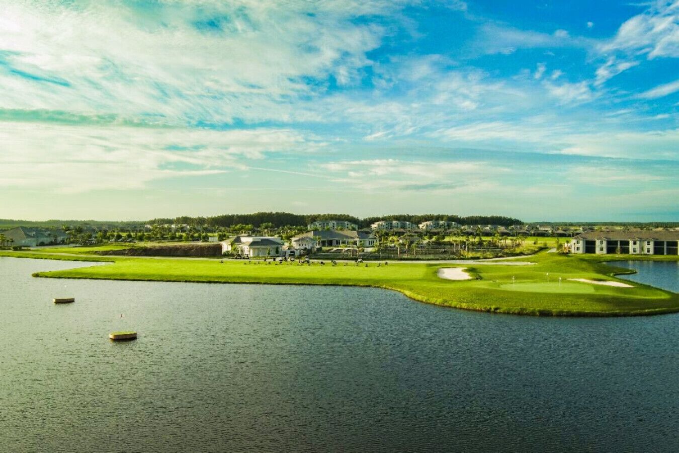 A golf course with a water body, clubhouse, and residential buildings in the background.
