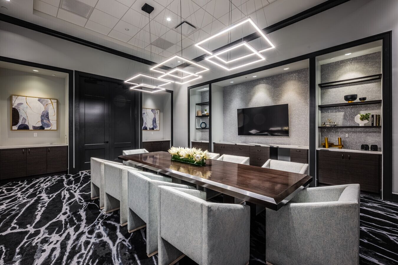 A modern conference room with a long table, grey upholstered chairs, a floral centerpiece, a flat-screen TV, and a decorative shelf with books and small plants.