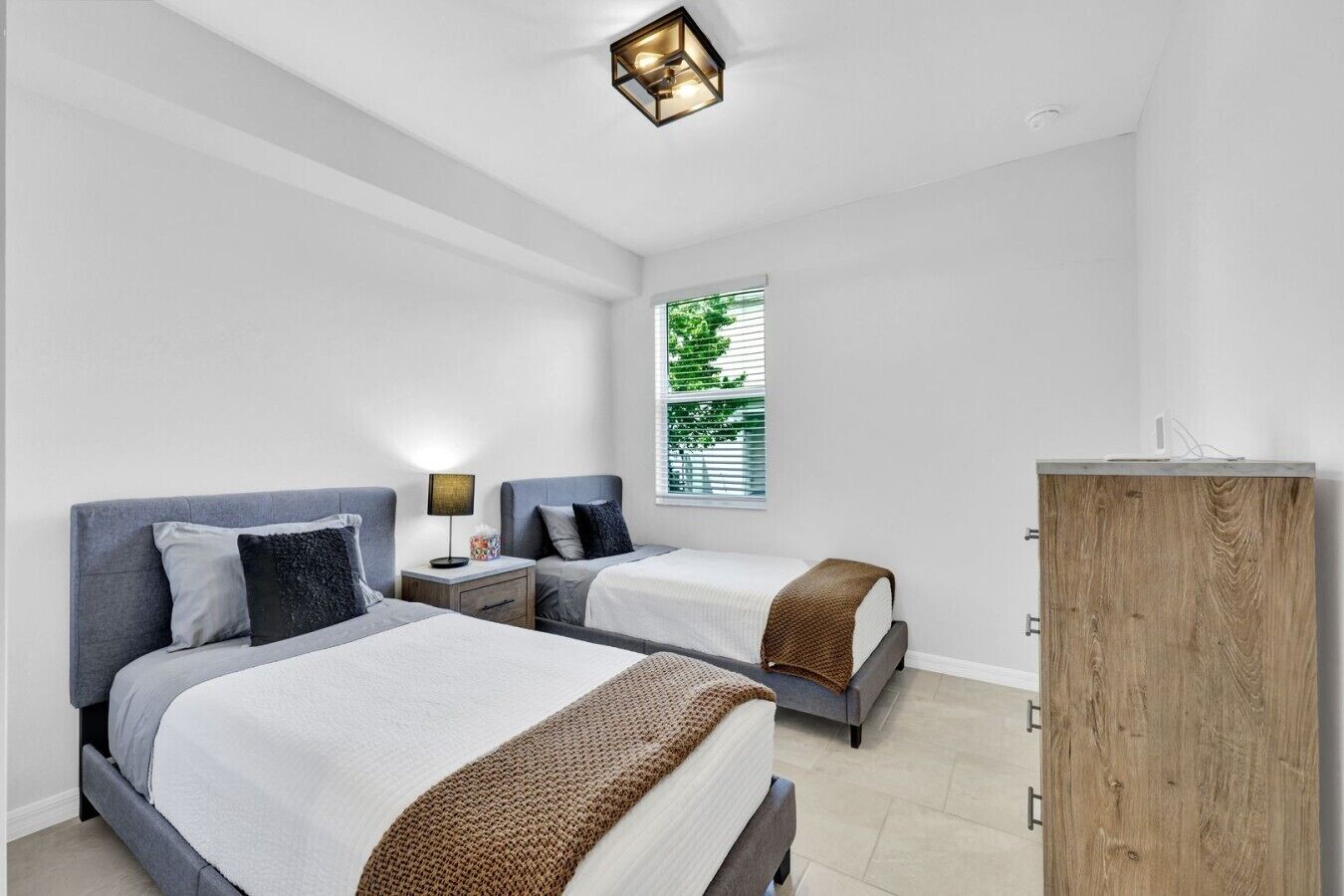 A modern bedroom with two beds, a wooden cabinet, and a window with blinds.