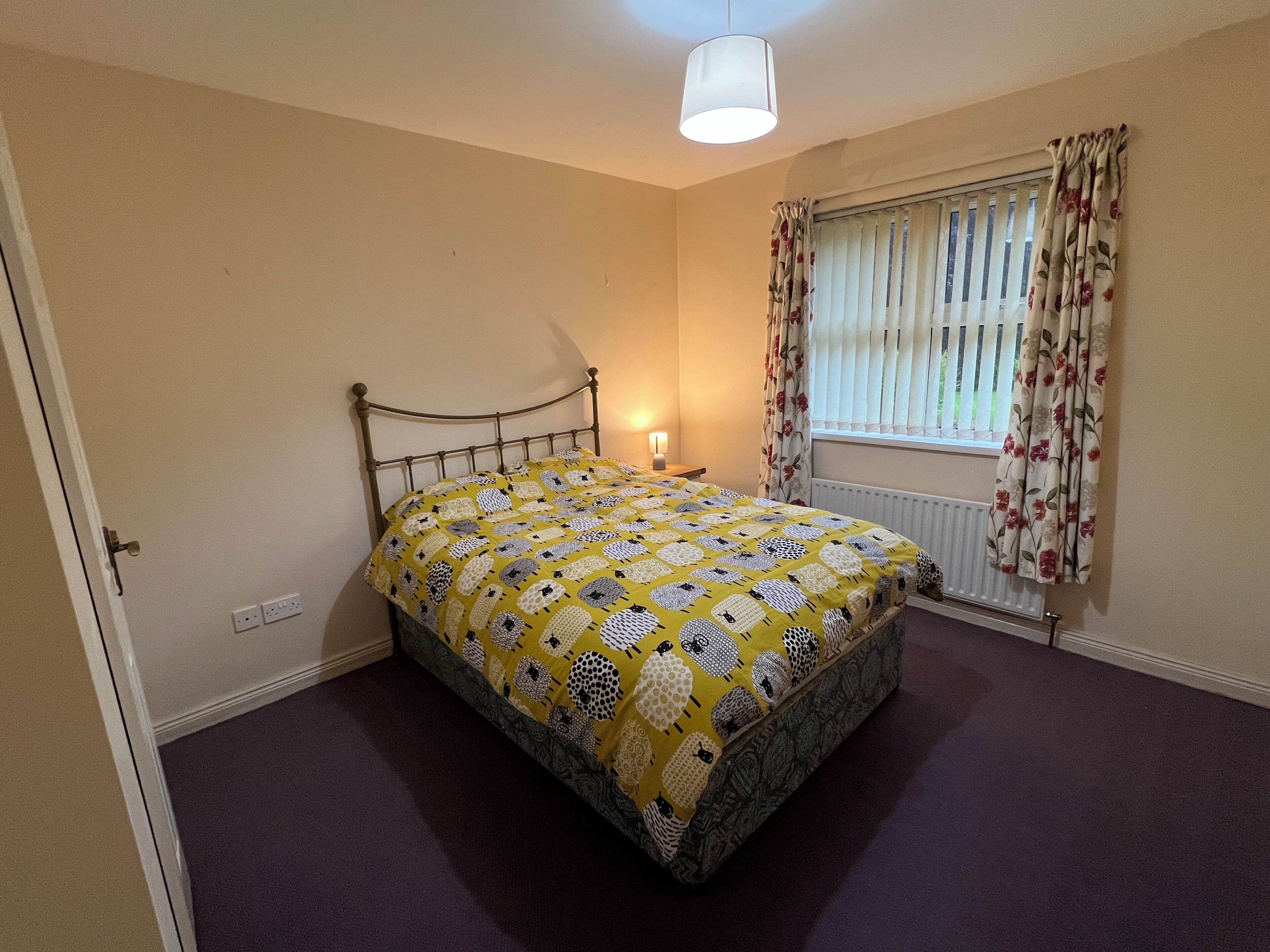 A bedroom with a bed, a window with curtains, a radiator, and a hanging light fixture.