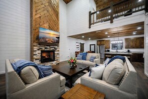 A spacious living area with a fireplace, a sofa set, a coffee table, and a kitchen area with a refrigerator and cabinets. - New Family Cabin/Treehouse/Hot Tub/EV Charger/Foosball/Corn Hole/Fire Pit (Broken Bow)