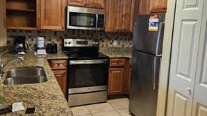 Una cocina con gabinetes de madera, electrodomésticos de acero inoxidable, incluyendo un microondas, horno y refrigerador, una encimera de granito y un salpicadero de azulejos.